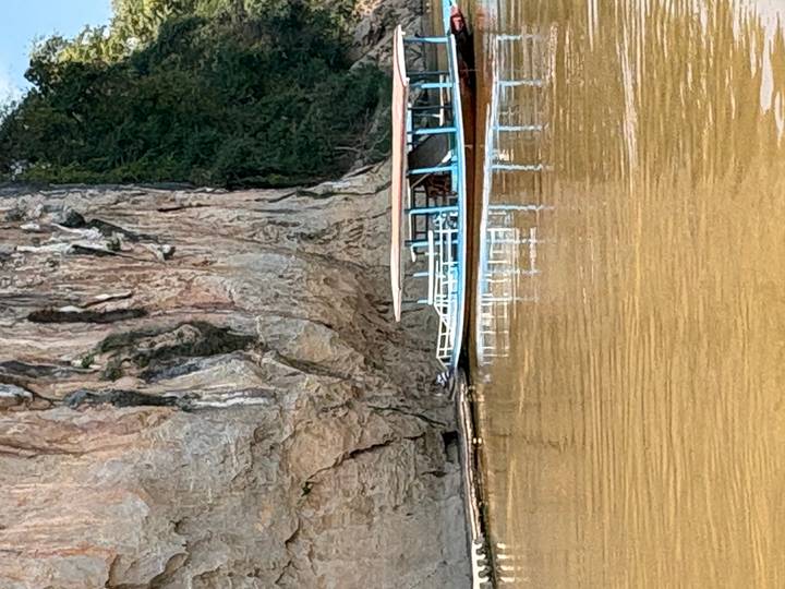 Long boat moored beside tall limestone cliff reflecting on brown river water.
