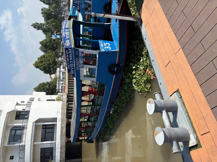 Blue riverboat docked beside riverside promenade with floating plants.