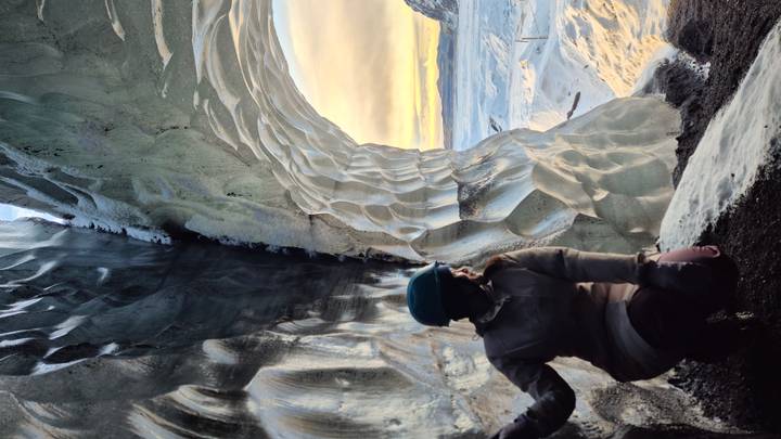 Explorer kneeling inside a luminously carved ice cave entrance overlooking snowy Icelandic plains at sunset