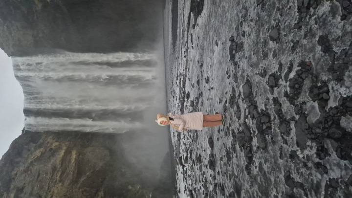 Person in winter coat standing on black gravel before the misty curtain of Skógafoss waterfall