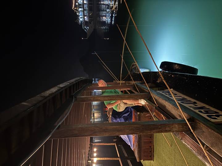 Man stepping off a wooden junk boat at night, green light illuminating the calm water in Halong Bay.