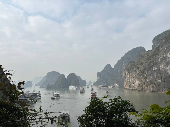 Wide view of Halong Bay dotted with cruise boats sailing between towering karst islands on a hazy day.