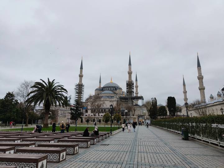 Visitors stroll in front of Istanbul’s Blue Mosque, partially covered by restoration scaffolding on a grey day.