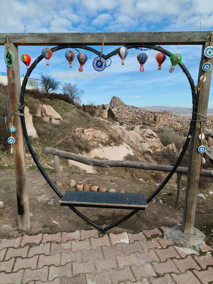 Scenic overlook with a large wooden swing frame decorated with Nazar evil-eye charms, looking out over Cappadocia’s rocky fairy-chimney landscape and Uchisar Castle.