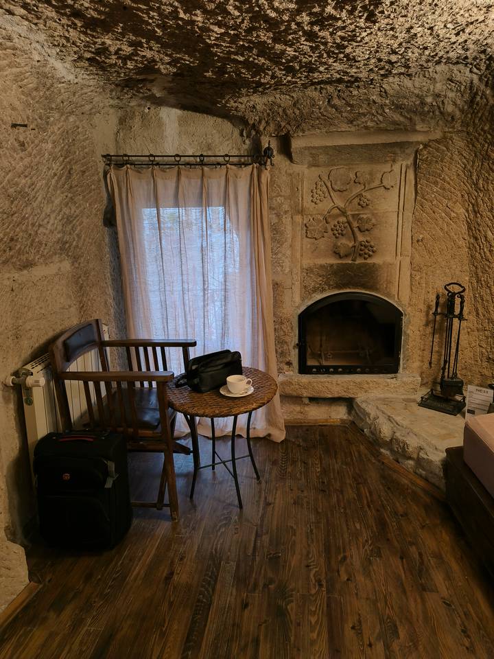 Cozy cave-style hotel room with stone fireplace, wooden chair, small table with a coffee cup and travel bags, softly lit by daylight through curtains.