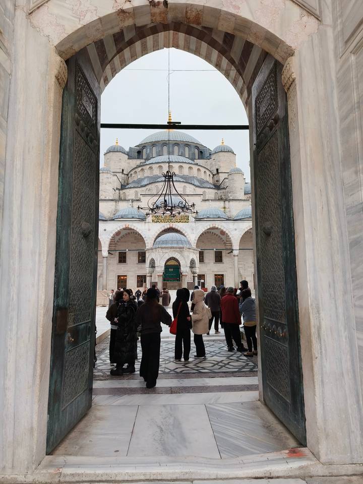 Visitors enter the grand marble courtyard of an Ottoman mosque through ornate bronze doors, framed by columns and arches.