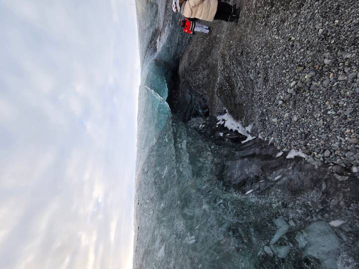 Jagged wall of blue glacial ice with gravel foreground under a cloudy Arctic sky.