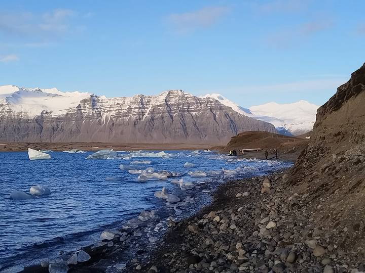 Shoreline of a glacier lagoon with scattered ice chunks and two people photographing the scene