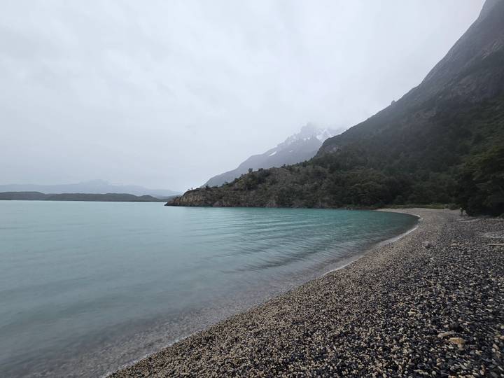 Overcast view of a tranquil turquoise lake and pebble beach beneath misty Patagonian peaks.