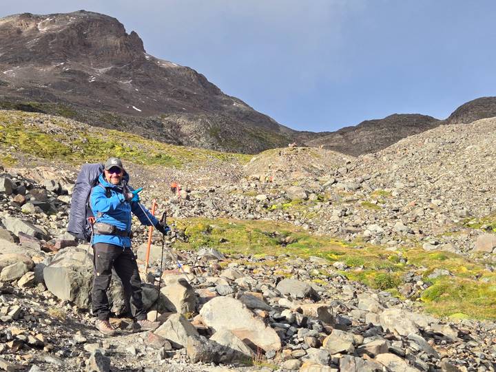 A solo trekker with a large backpack and trekking poles makes their way across a rocky mountain slope under clear skies.