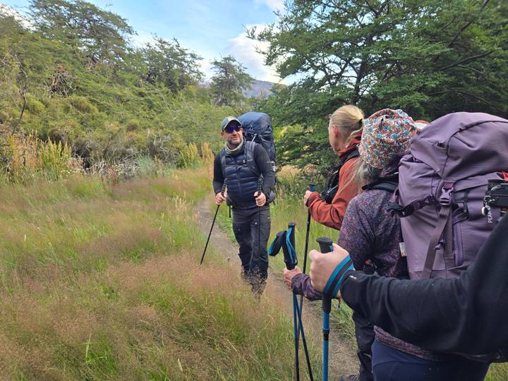 Hikers carrying backpacks and trekking poles chat while walking along a grassy trail lined with shrubs and trees.
