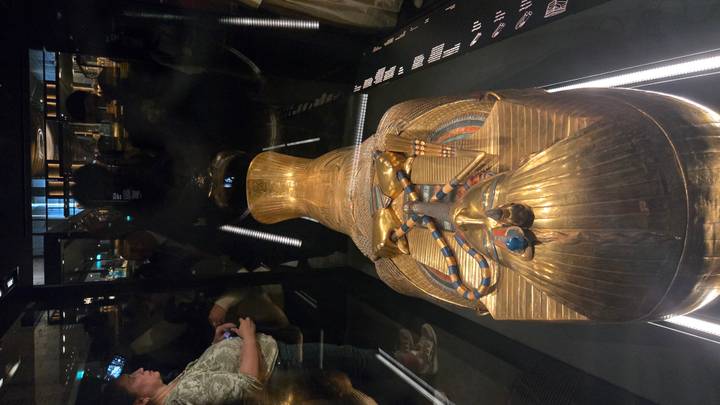 A golden sarcophagus displayed in a dimly lit museum case with visitors reflected in the glass.