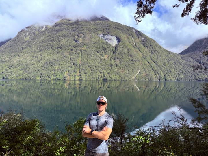 A man wearing sunglasses poses in front of a still lake that perfectly mirrors a lush green mountain under patchy clouds.