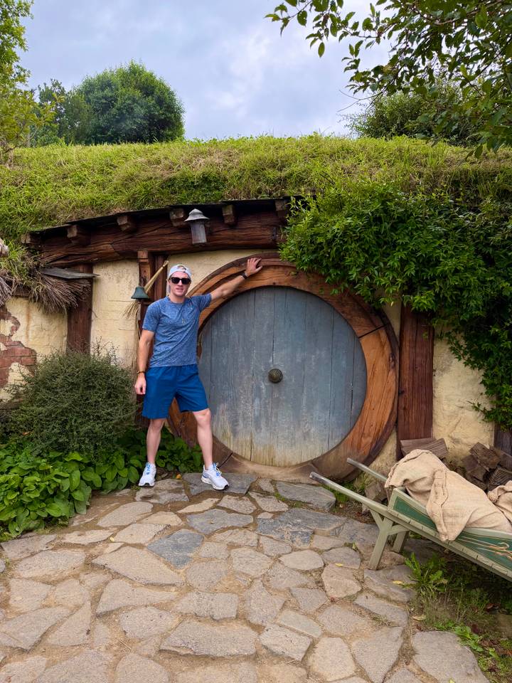 A visitor smiles beside the iconic round wooden door of a Hobbiton house set in lush greenery.