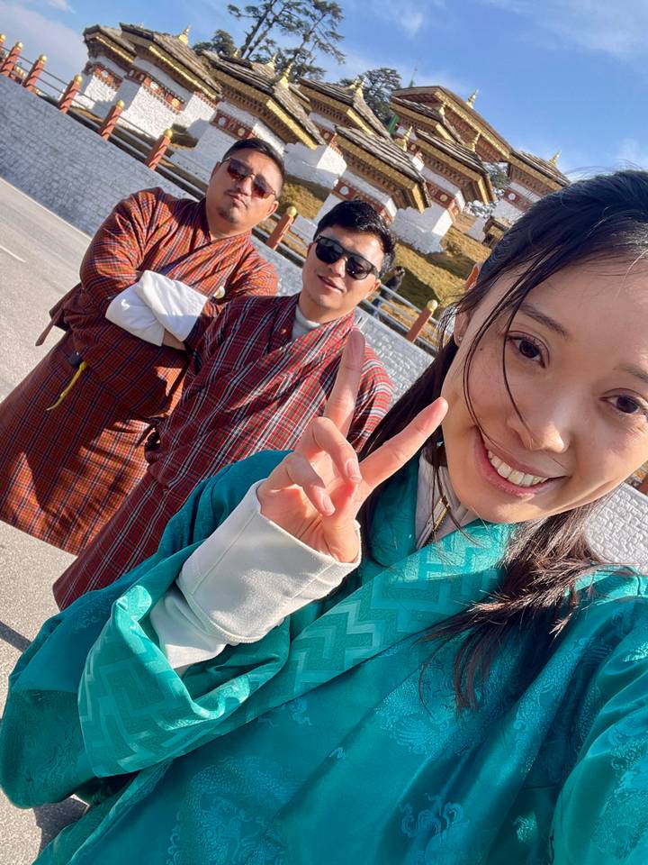 Close-up selfie of a smiling woman making a peace sign with two friends in Bhutanese robes behind her.