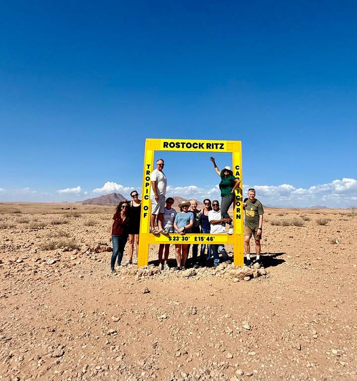Travel group poses around a bright yellow Tropic of Capricorn frame in a vast Namib desert landscape.