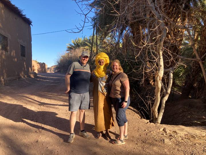 Two tourists pose with a local man in yellow turban on a dusty palm-lined village track.