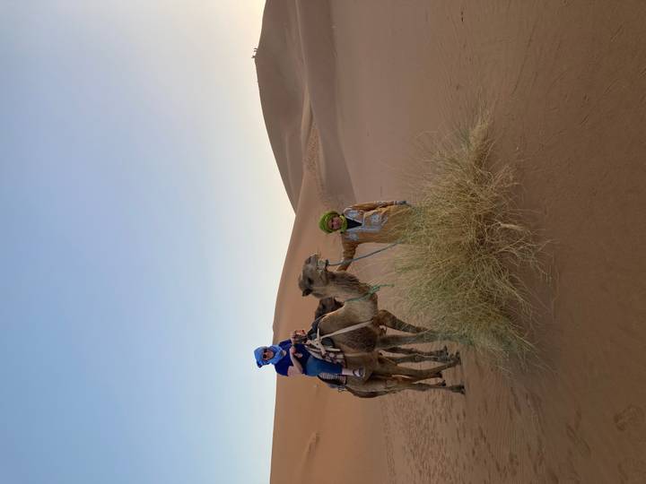A tourist on camelback and a guide stand amid sweeping Sahara sand dunes in soft evening light.