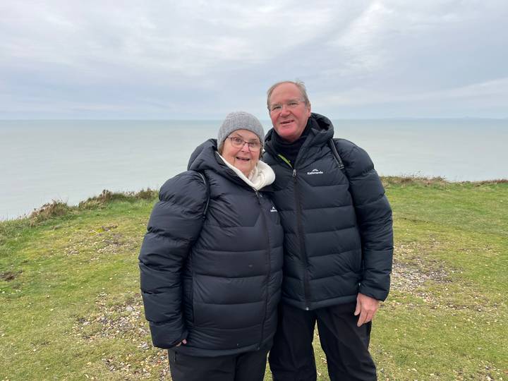 Smiling older couple in puffer jackets stand on a grassy cliff top with grey sea beyond.