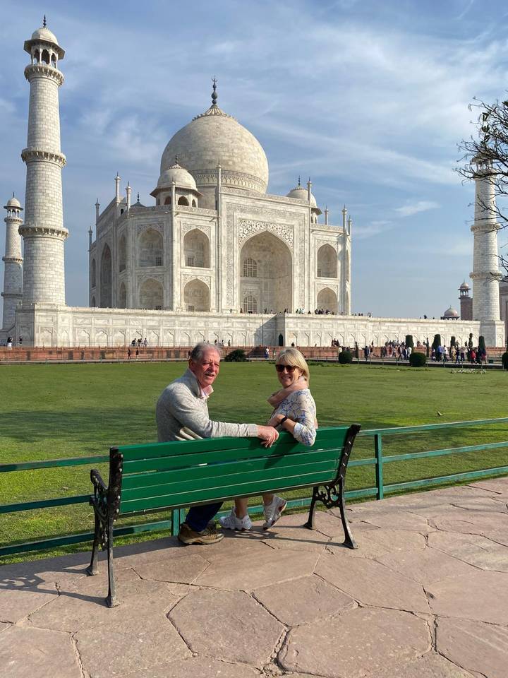 Couple seated on a green bench happily pose with the majestic Taj Mahal rising behind them on a sunny afternoon.
