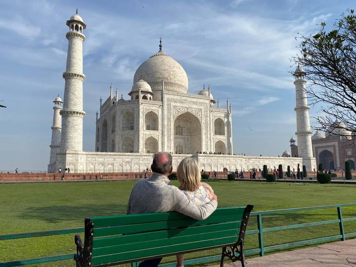 Couple sits arm-in-arm on a bench gazing at the Taj Mahal across green lawns under a clear blue sky.