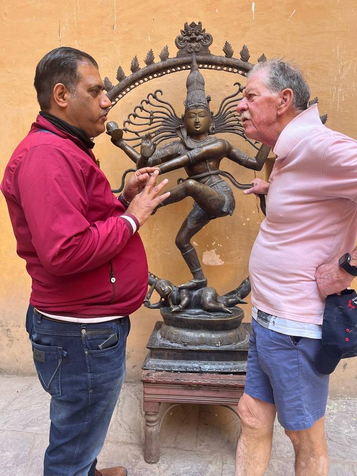 Two men chat animatedly beside a bronze dancing Shiva statue displayed against a yellow courtyard wall.