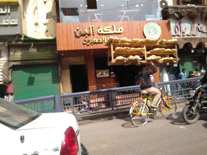 Cyclist balances towering trays of bread on head while navigating a busy Cairo street.