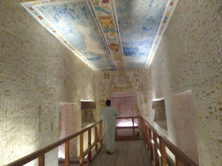 Dimly lit corridor inside an ancient Egyptian tomb covered with hieroglyphic carvings, a man in traditional robe walks along a wooden walkway toward a decorated chamber.