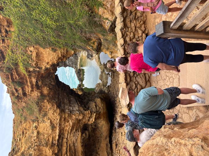 Travellers descend stairs toward a natural rock arch and tidal pool set in golden cliffs.