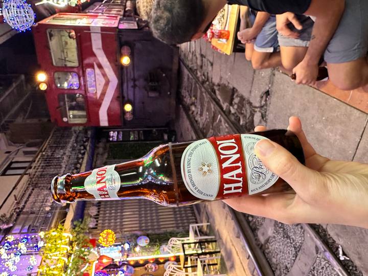 Hand holding a Hanoi beer bottle with a red locomotive approaching on train street at night.