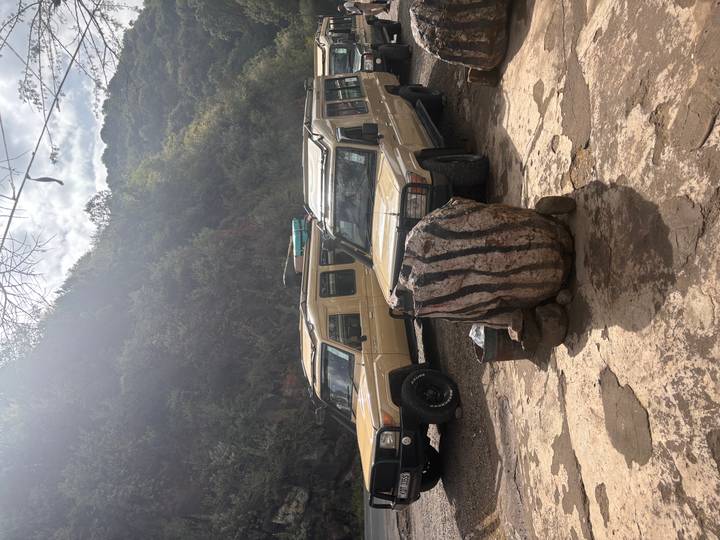 Two beige safari jeeps parked on a rough concrete forecourt beside a rocky hill.