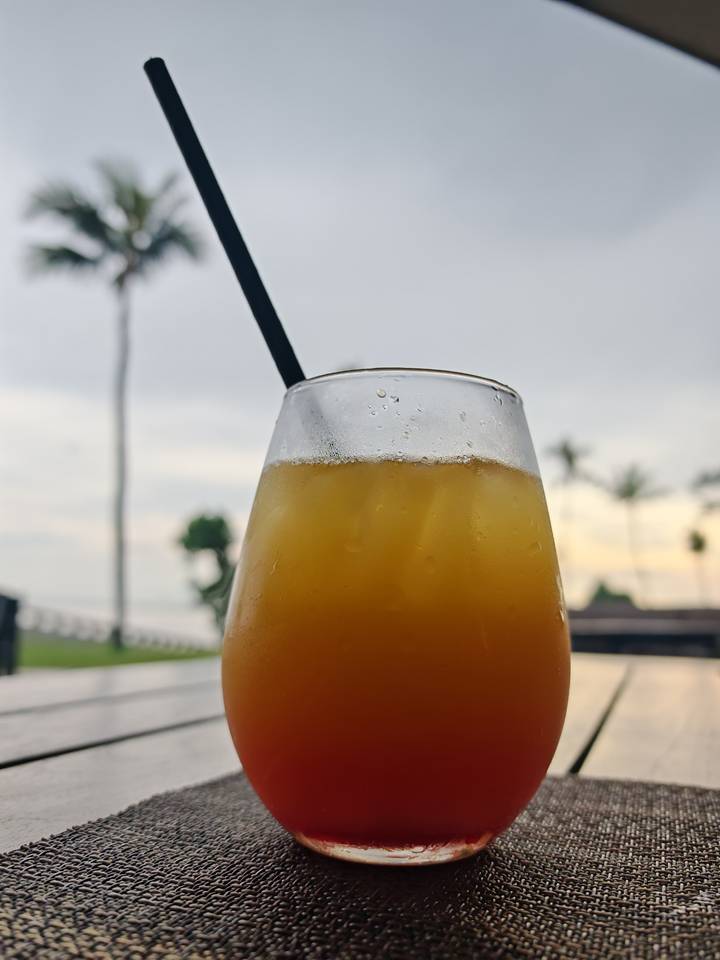 Close-up of an iced tropical drink in a glass with blurred palm trees and ocean backdrop.