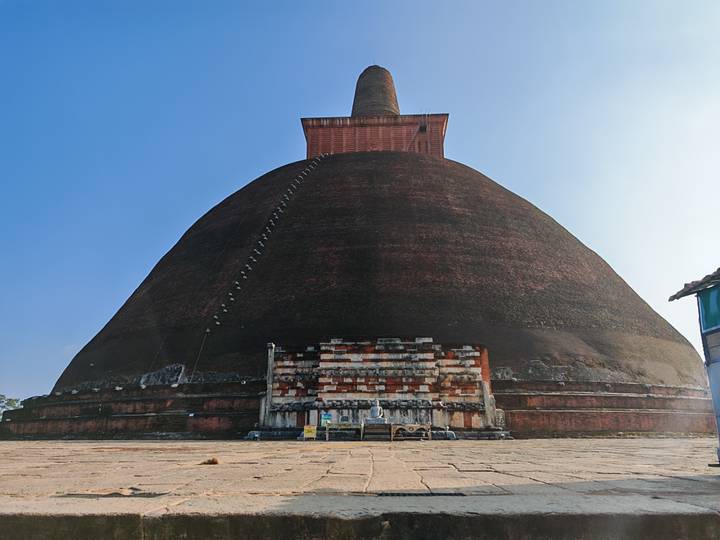 Massive brick stupa rising toward a clear blue sky with a small entrance base, photographed from ground level.