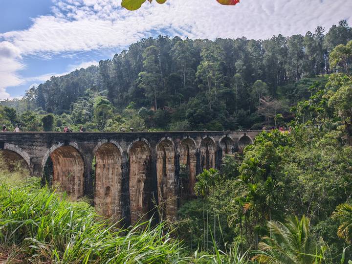 Historic Nine Arch railway bridge spans a lush jungle gorge with small tourists walking along its top.