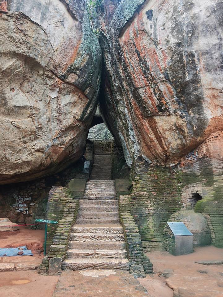 Stone staircase leads through a narrow cleft between massive weather-worn boulders with brick walls below.