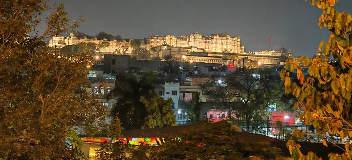 Nighttime cityscape of a lit hilltop palace above treetops and colorful houses.