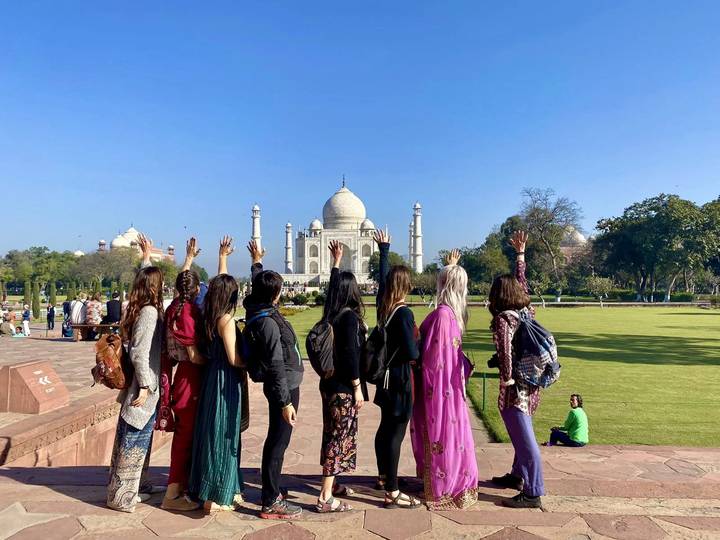 Group of travelers raise hands toward the Taj Mahal on a sunny morning.