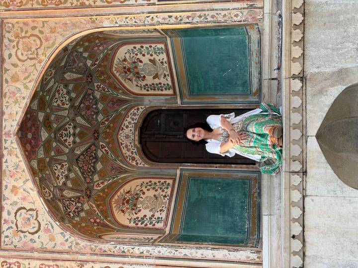 Woman sits in a richly painted Mughal archway surrounded by floral murals.