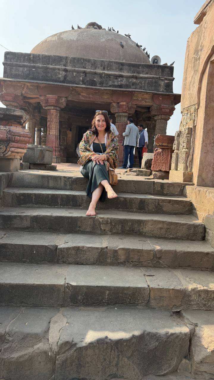 A barefoot woman sits casually on worn stone steps of an old sandstone temple or fort.