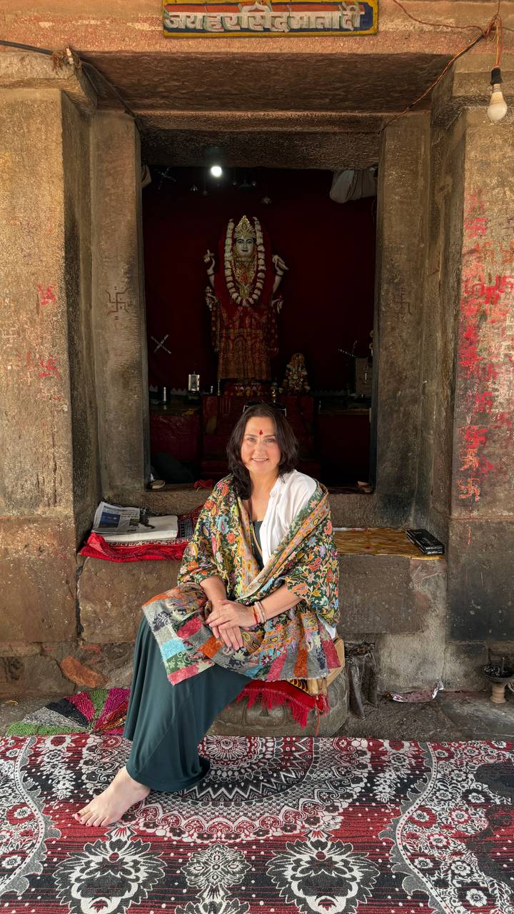 Woman with a red bindi smiles while seated in the entrance of a dimly lit shrine.