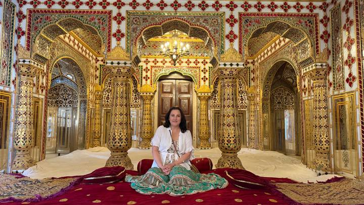 Woman sits inside a dazzling mirrored and gilded chamber adorned with intricate Rajasthani decor.