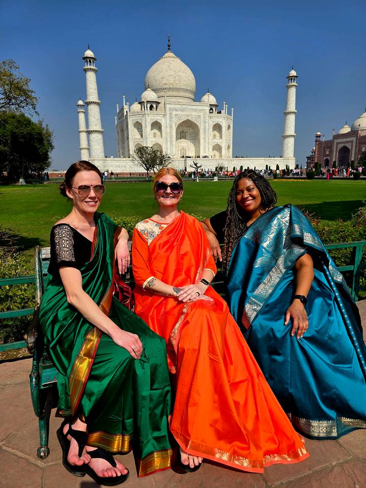 Three smiling women in vibrant saris sitting on a bench with the Taj Mahal rising behind them