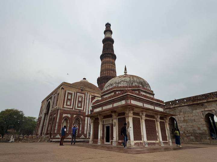 View of Qutub Minar and adjacent domed mausoleum with tourists at the base on an overcast day