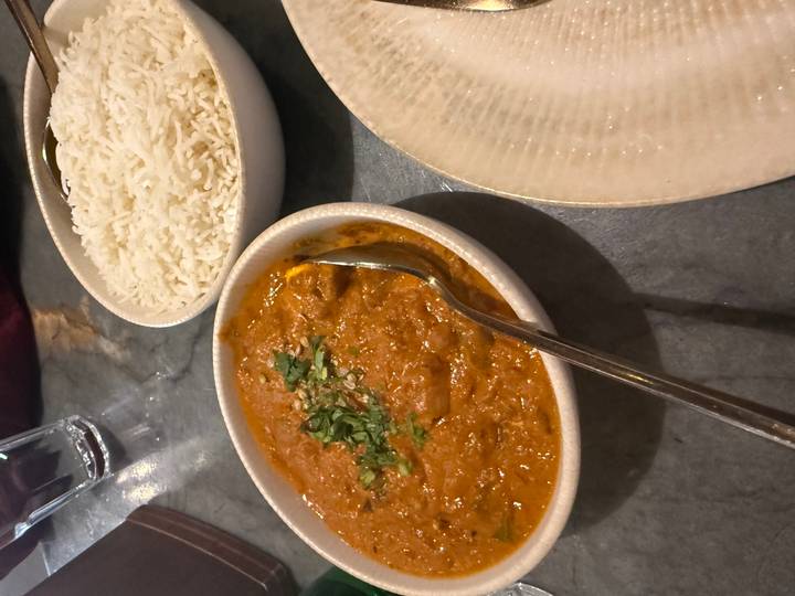 Close-up of a bowl of rich curry garnished with herbs alongside steamed basmati rice on a stone table