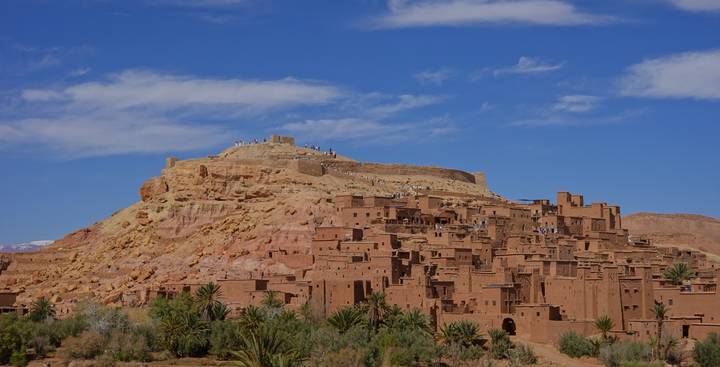 Clay-colored buildings of Ait Benhaddou climb a rocky hilltop beneath a bright blue Moroccan sky.