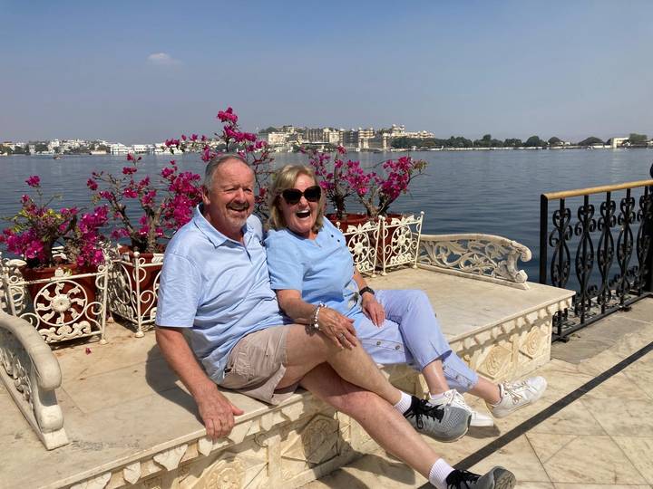 Laughing couple seated on a marble bench overlooking Lake Pichola and Udaipur’s city palace.