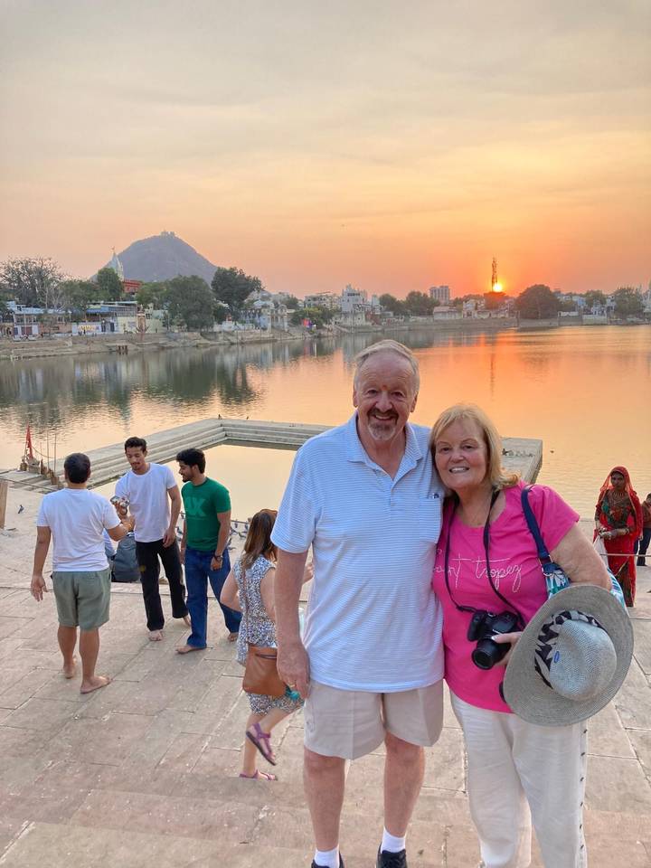 Couple at Pushkar Lake during sunset with temple steps and orange sky reflected in the water.