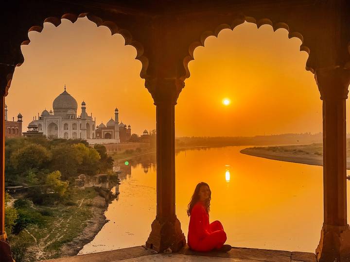 Woman in red looks across the Yamuna River to the Taj Mahal at sunrise, framed by ornate arch columns