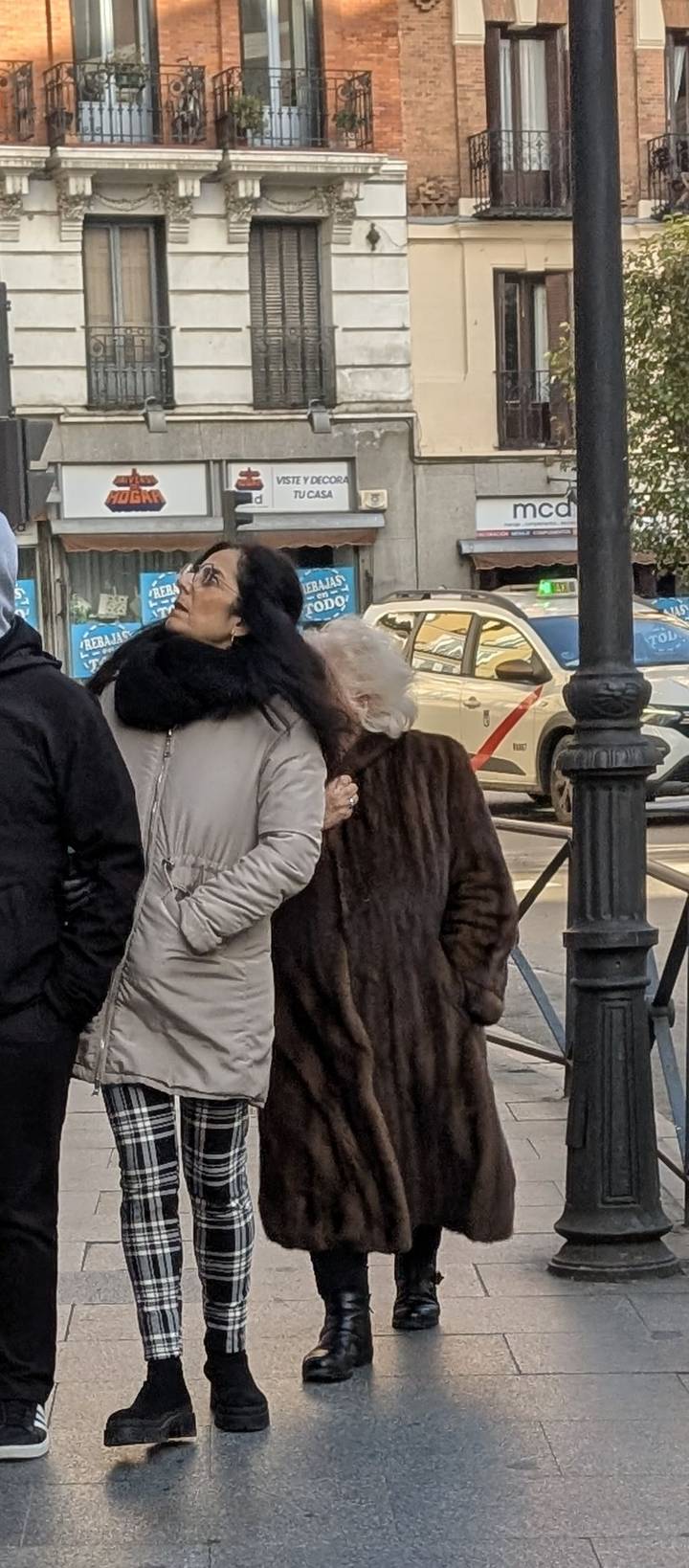 Older and younger women bundled in coats wait at a city crossing.