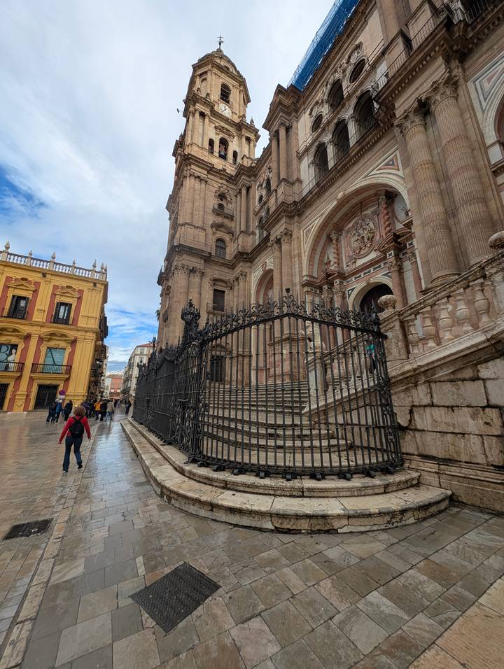 Rain-washed street beside Malaga Cathedral with pedestrians and historic buildings.
